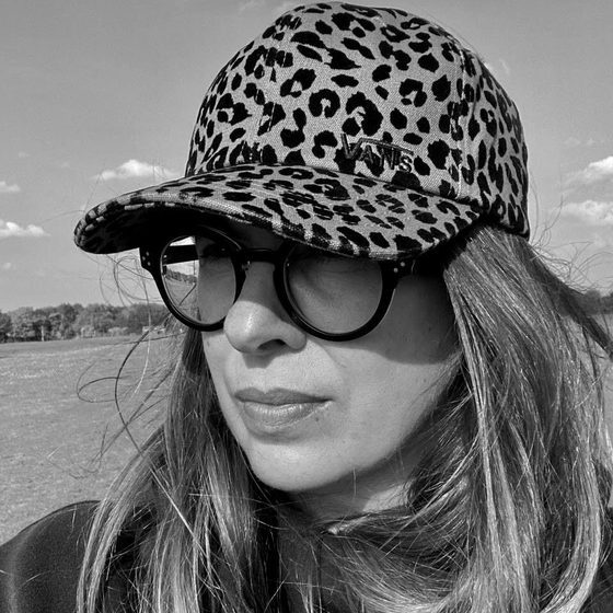 Sarah Dennehy, a woman with light-skin and long hair, is wearing a leopard print baseball cap and black glasses. The image is in black and white.