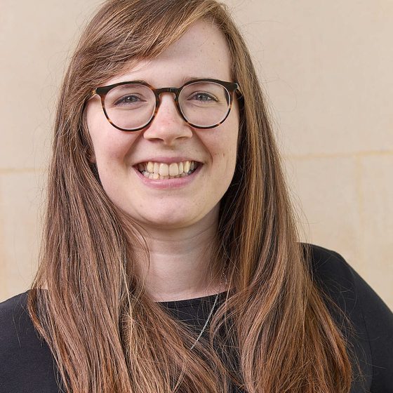 Esther Poole, a woman with light skin tone, long dark blonde hair. She wears a black top, tortoiseshell glasses and a wide, friendly smile.